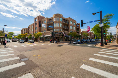 An urban intersection with a brick apartment building, cars, and pedestrians under a partly cloudy sky.