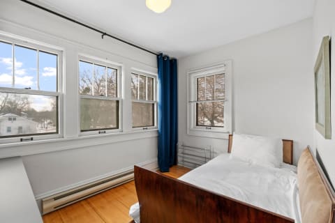 A cozy bedroom featuring a single bed, large windows with blue curtains, and wooden flooring.