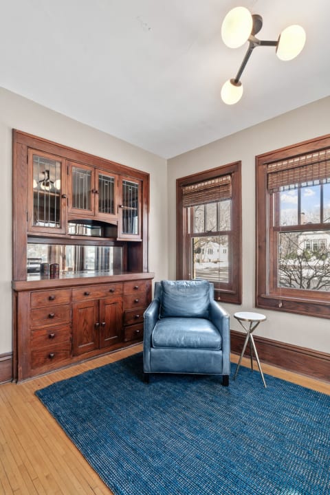 A stylish living room corner featuring a blue leather armchair, wooden cabinet, and large windows.