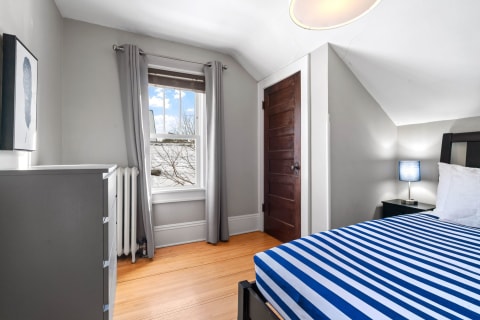 A well-lit bedroom featuring a bed with a striped mattress, a gray dresser, and a window with curtains.