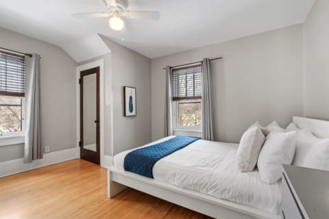 A cozy bedroom featuring a white bed with blue accents, wooden flooring, and natural light from the window.