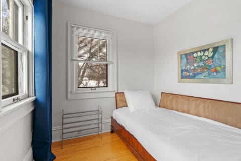 A minimalist bedroom corner with a wooden bed, blue curtain, and framed still life artwork.