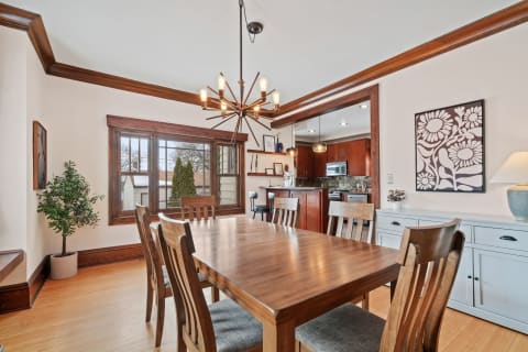 A modern dining room featuring a wooden table, chairs, potted plant, chandelier, and views into a kitchen.