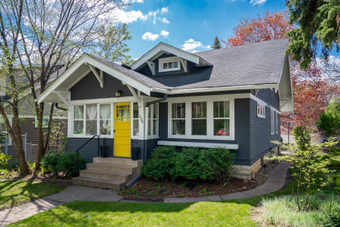 A gray house with a yellow front door, surrounded by green plants and clear blue sky.
