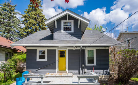 A gray house with a yellow door and blue chairs on a wooden deck under a blue sky with clouds.