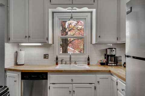 A modern kitchen with white cabinetry, a wooden countertop, and a window overlooking a fall tree.