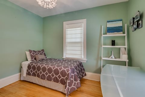 A single bed with burgundy and white patterned bedding in a light green room, featuring a shelf with decorative items and a white desk.
