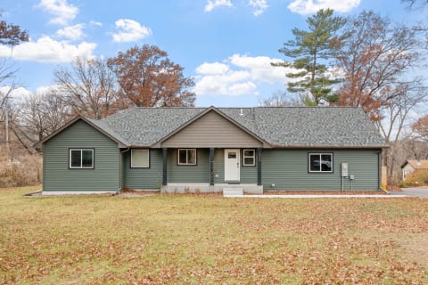 A green single-story house with a large yard covered in fallen leaves under a blue sky.