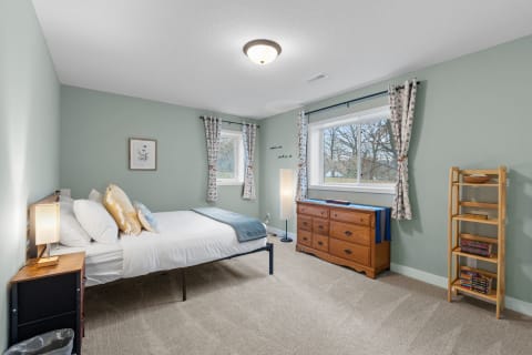 A serene bedroom featuring a bed with decorative pillows, a dresser, and a window with floral curtains.