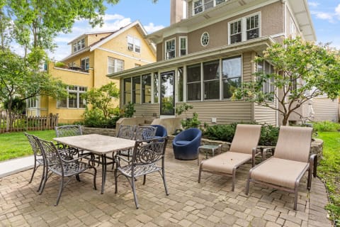 A private patio featuring a stone dining table, metal chairs, and lounge chairs surrounded by greenery and house background.