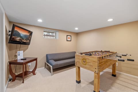A cozy game room with a foosball table, a gray couch, and a mounted TV displaying a scenic landscape.