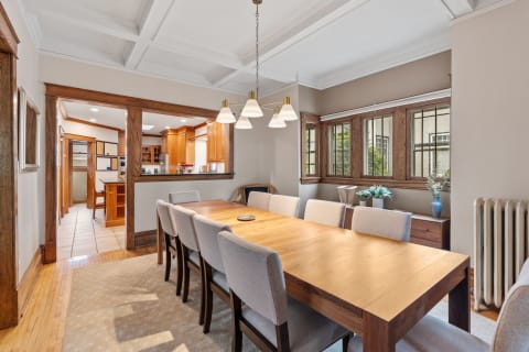 A spacious dining room featuring a long wooden table, upholstered chairs, and a modern pendant light, with a view into the kitchen through an archway.