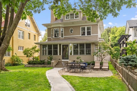 Two-story house with wraparound porch, patio seating area, and surrounding greenery.