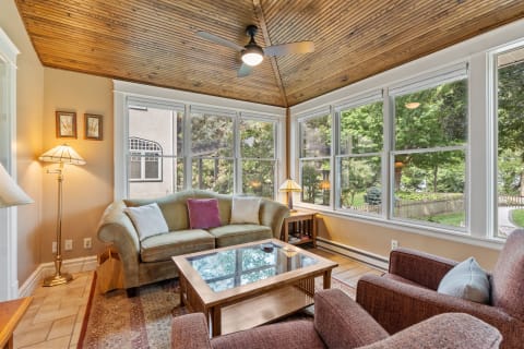 Cozy sunroom with large windows, olive green sofa, and wooden ceiling overlooking a garden.