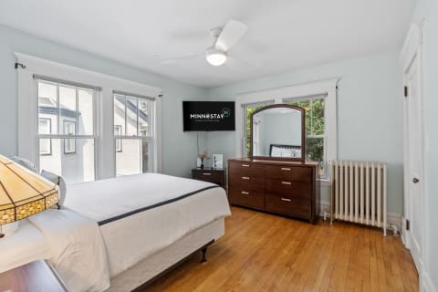 A serene bedroom with hardwood floors, a ceiling fan, and large windows, featuring a bed, dresser, and television.