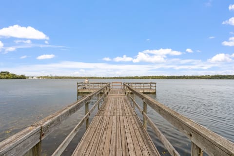 A wooden dock extending over a calm lake with a person fishing in the background and blue sky above.