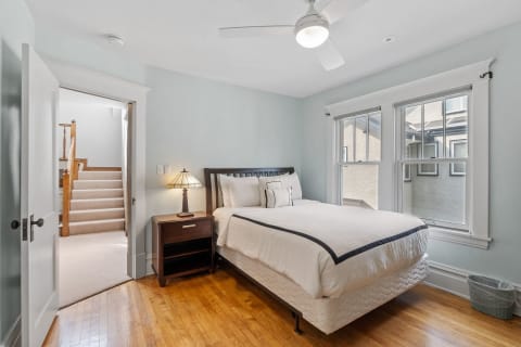 A peaceful bedroom featuring blue walls, hardwood floors, a queen-sized bed, and a staircase in the background.