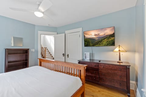 A peaceful bedroom with blue walls, wooden furniture, a flat-screen TV displaying mountains, and a ceiling fan.