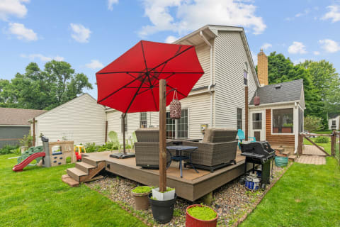 A wooden backyard deck with wicker chairs, a red umbrella, and children's play equipment.