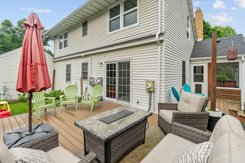 A backyard patio with wicker furniture and a red umbrella, surrounded by greenery.