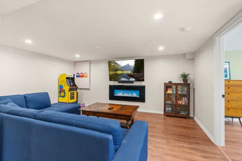 A stylish basement lounge area with a blue sofa, arcade game, TV, and fireplace.