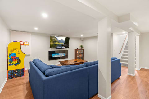 A modern basement featuring a blue sectional sofa, an arcade game, a mounted TV, and an electric fireplace.