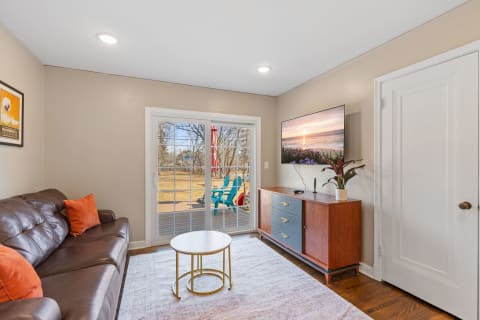 A cozy living room featuring a brown leather sofa, a round coffee table, and a mounted TV, with sliding doors leading to a deck and outdoor seating.
