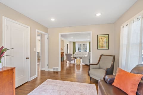 A cozy living room with beige walls, a brown leather sofa, grey armchair, and a glimpse of a dining area beyond.