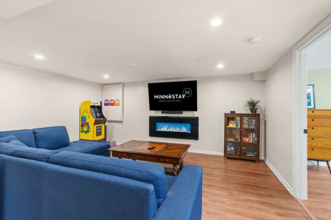 Modern basement living area featuring a blue sectional sofa, vintage arcade game, and wall-mounted television with an electric fireplace.