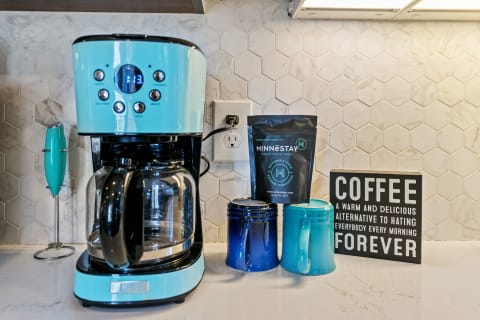 A vibrant kitchen counter with a turquoise coffee maker, mugs, and a humorous coffee sign.
