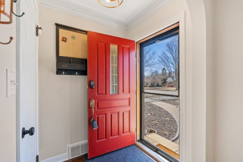 An open red front door leading into a bright hallway with hooks and a corkboard.