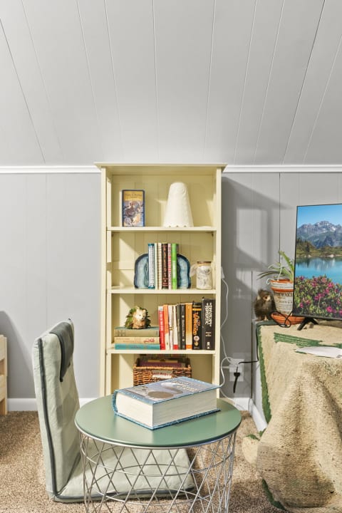A cozy room corner with a yellow bookshelf filled with books, a green table, and a television showing a mountain scene.