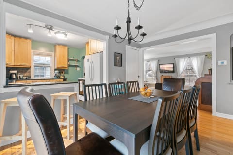 A dining room with a wooden table, surrounded by chairs, leading into a kitchen with green walls and wooden cabinets.