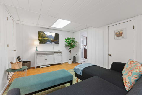 A cozy modern living room featuring a dark gray sofa, decorative pillows, a teal ottoman, and a landscape TV display.
