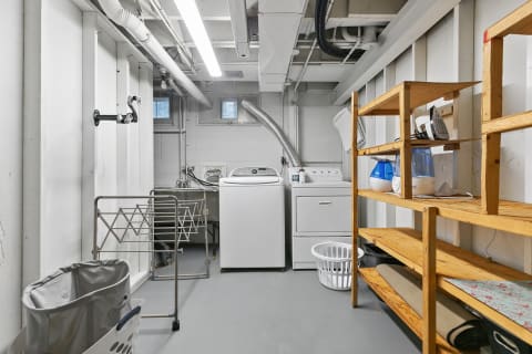 A spacious laundry area in a basement featuring washing and drying machines with shelving and drying racks.