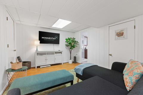 A cozy living area with dark gray sofa, blue ottoman, and wall-mounted TV displaying MINNÊSTAY logo.