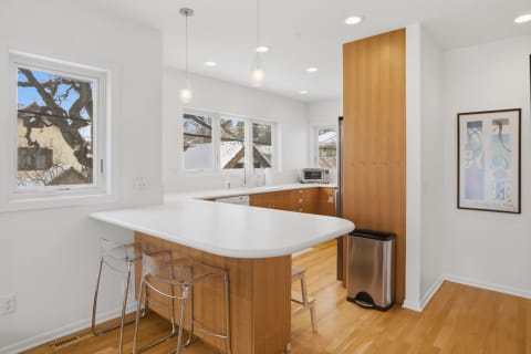 Modern kitchen with white countertops and warm wood cabinetry, illuminated by natural light through large windows.