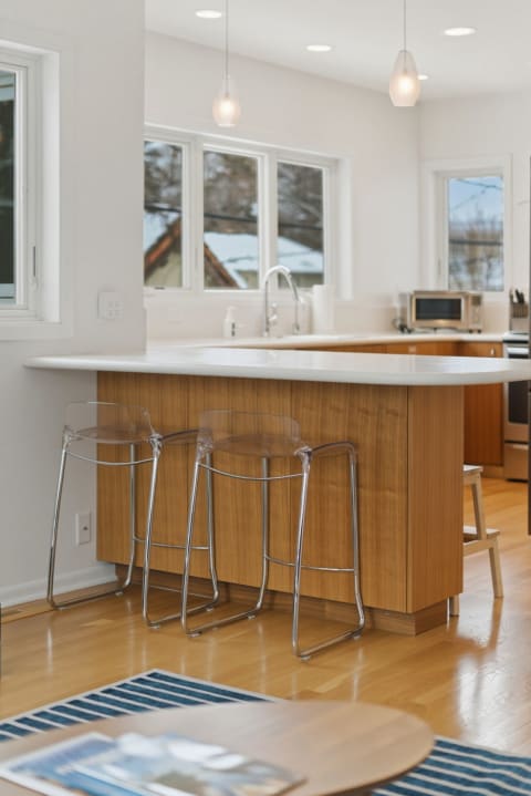 A modern kitchen with a wooden island, white countertop, and transparent bar stools, lit by natural light from large windows.