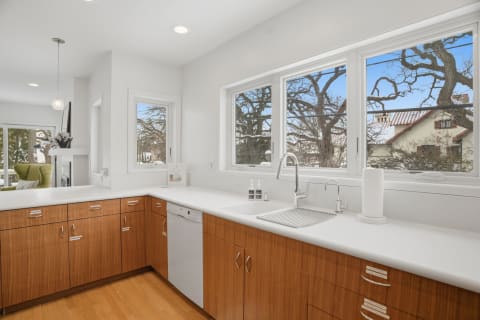 Modern kitchen with white countertops and wood cabinetry, showing large windows and natural light.