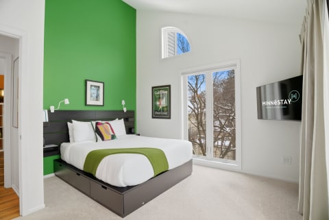 Modern bedroom featuring a bright green wall, white bedding, and windows with a view of trees outside.