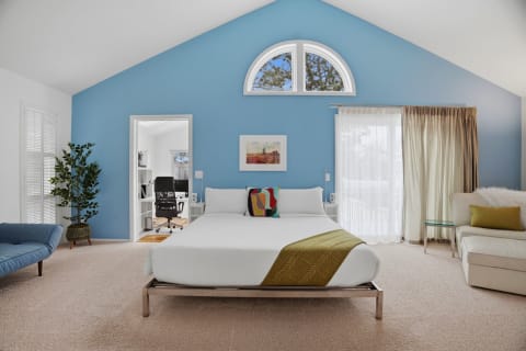 A modern bedroom with vaulted ceilings, featuring a white bed with colorful pillows, a blue chair, and a workspace.