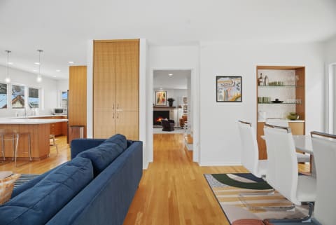 An open-concept living area showcasing a navy blue sofa, white dining chairs, and a warm wood kitchen, with a glimpse of a fireplace in the background.