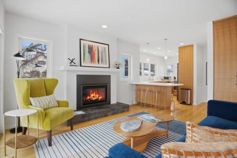 Bright living room with a fireplace, colorful armchair, and modern kitchen in the background.
