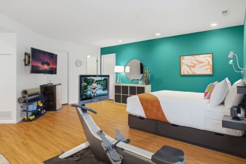 Modern bedroom with teal accent wall, bed with orange throw, rowing machine, and wall-mounted TV.