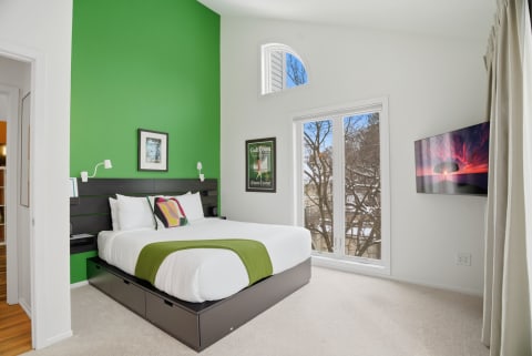 A vibrant bedroom featuring a green accent wall, a bed with white linens, and a large window with a view of trees outside.