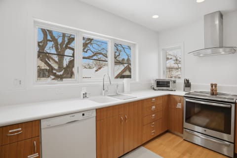 A bright modern kitchen featuring white countertops and wooden cabinetry, with large windows overlooking a tree.