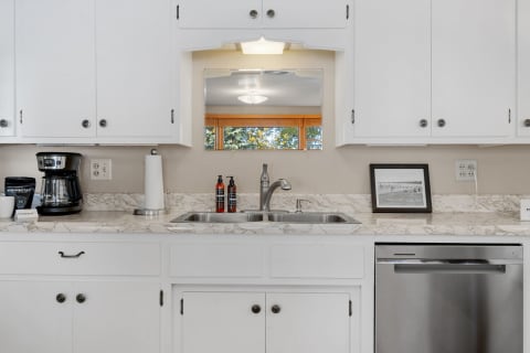 A kitchen featuring a stainless steel sink, coffee maker, and marble countertops under white cabinets.