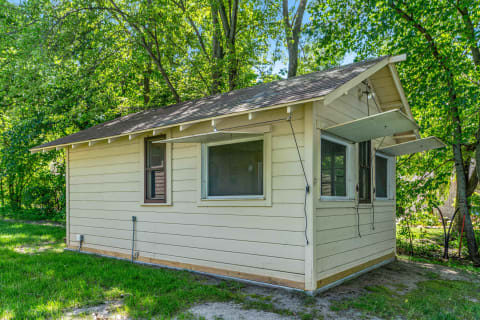 A light beige wooden cottage with an open awning window, nestled among green trees.