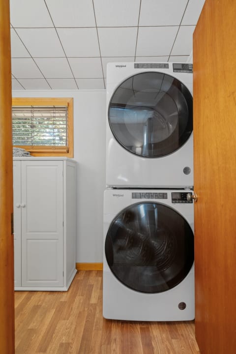Compact laundry room featuring stacked white Whirlpool washer and dryer next to a white cabinet and a window.