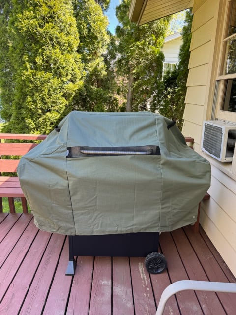 A green cover obscures a black grill on a wooden deck with evergreen trees in the background.
